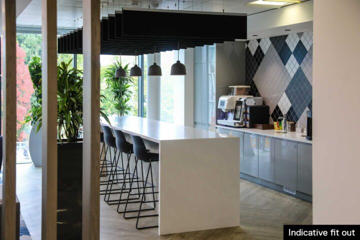 Modern office kitchen with high white counter, bar stools, plants, pendant lights, and coffee area.