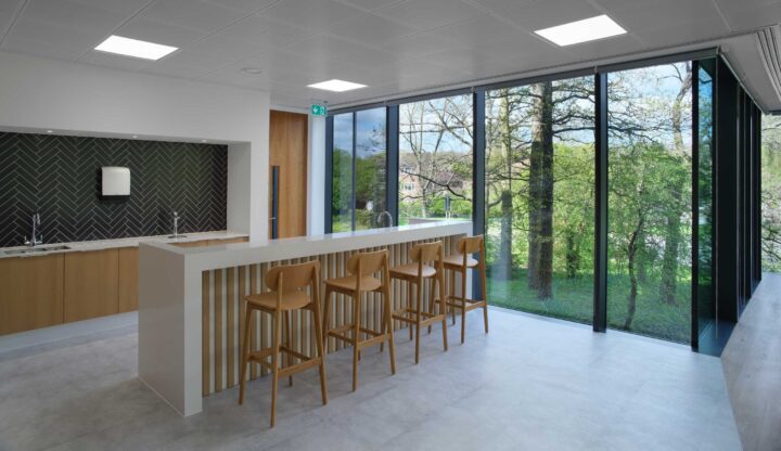 Modern kitchenette with a white island and four wooden stools, large windows with a view of trees and a building, black backsplash, and overhead lights.