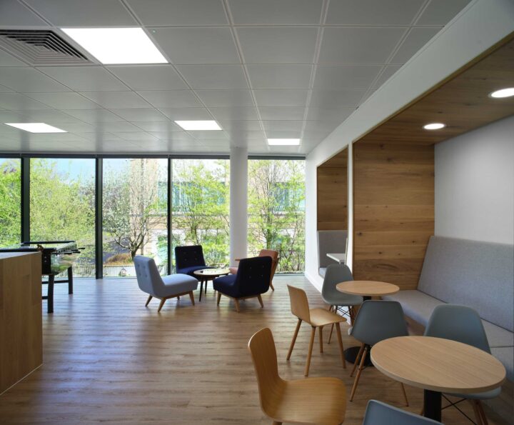 A modern office lounge with wooden floors, various chairs, tables, and a booth seating area. Large windows provide natural light and a view of trees outside.