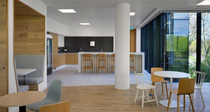 A modern office kitchenette and dining area with light wood furniture, gray bench seating, and large windows overlooking a tree-lined view.