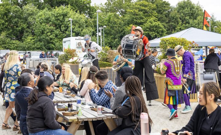 A diverse group of people is sitting at picnic tables enjoying food while a band in colorful outfits performs in the background at an outdoor event.