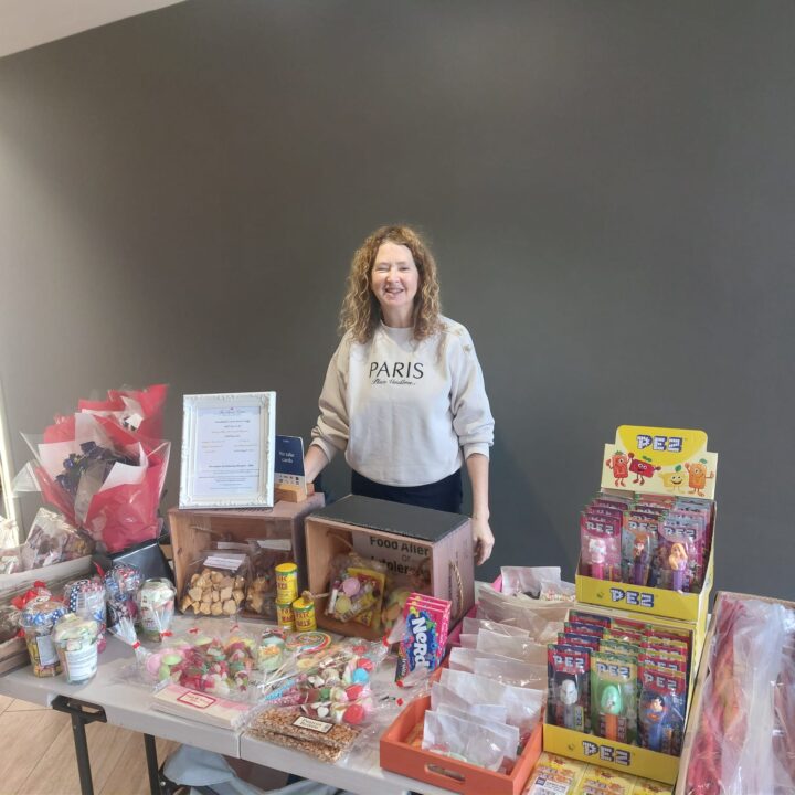 Woman stands behind a table with assorted snacks, treats, and candy arranged for sale.