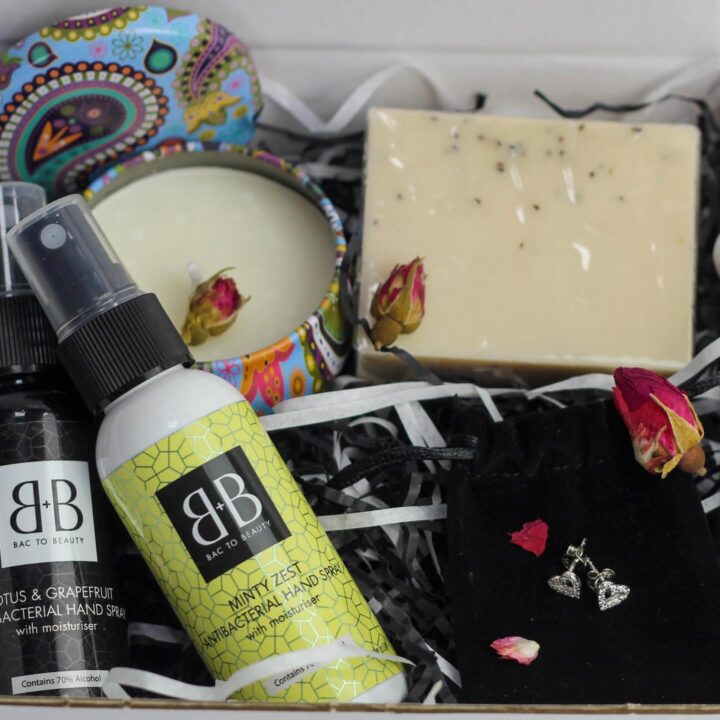 A gift box with hand sprays, a candle, soap, a pouch with earrings, and dried rosebuds.