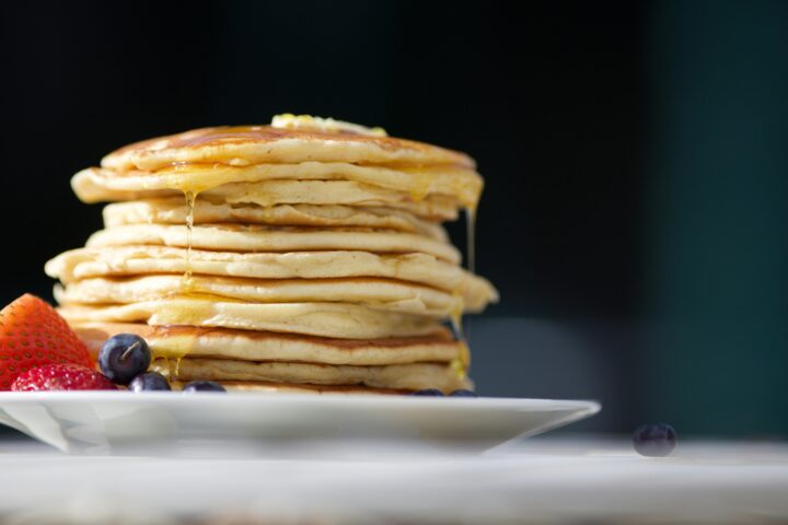 Stack of pancakes with syrup and a side of strawberries and blueberries on a white plate.