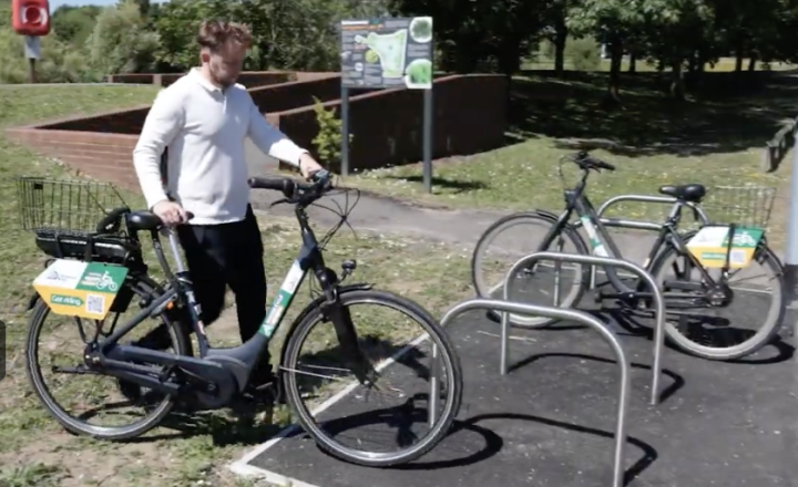 A person walks a rental bike toward a bike rack in an outdoor park area with greenery.